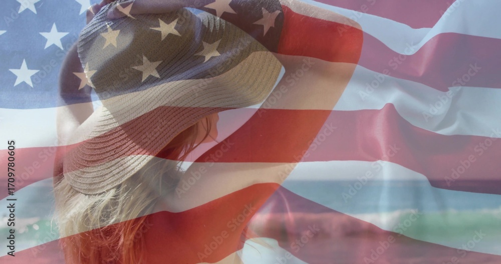 Fototapeta premium Raising bikini-clad female adjusting wide-brimmed sun hat on shore with ocean waves, flag overlay
