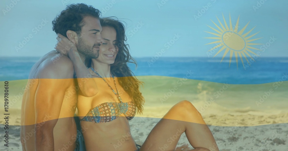 Fototapeta premium Embracing couple wearing swim trunks and bikini top sitting on calm beach, with flag overlay