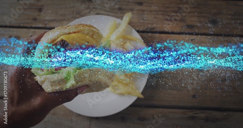 Lifting sesame-seed bun burger with adult hand over wooden tabletop, with ceramic plate of fries