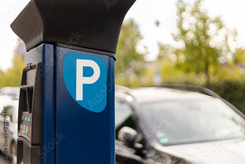 A parking meter is prominently displayed in a clean parking lot surrounded by greenery. Several cars are parked nearby, suggesting a bustling area during the day