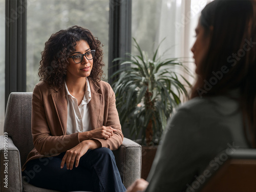A professional woman with curly hair and glasses, dressed in a brown blazer and white shirt, sits in an armchair attentively listening to another person in a modern setting