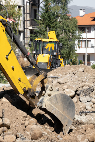 This image captures a yellow backhoe loader, a piece of heavy construction equipment, digging into rocky soil. The scene conveys ongoing construction or excavation work under a clear sky.