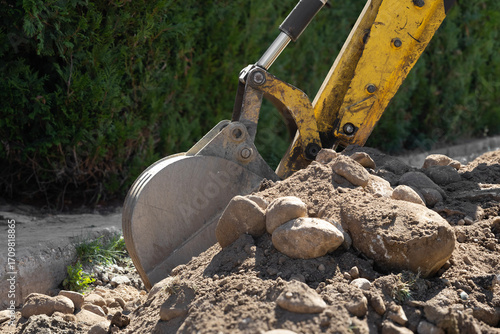 Heavy-duty excavator bucket digging earth and rocks. Powerful yellow arm against green foliage, showing active construction or landscaping work.