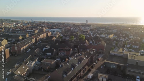 Aerial view of a coastal town at sunset with a white church, red-roofed houses, grassy dunes, and beach cabanas. The golden sky and calm ocean create a serene, picturesque atmosphere.