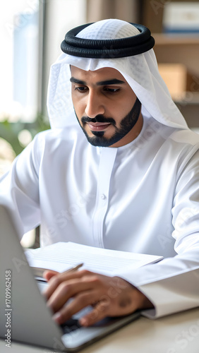 Focused Emirati businessman working on laptop in a modern office setting.