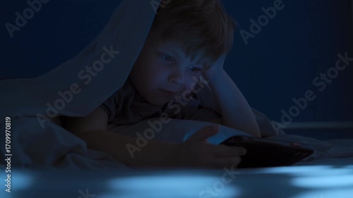 Contented little boy lies in bed at night under the blanket, holding his phone, illuminated with blue light of screen, while watching videos or cartoons. Screen time gadget overuse among children