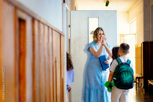 Smiling teacher welcomes a group of school kids in front of the classroom at hallway.