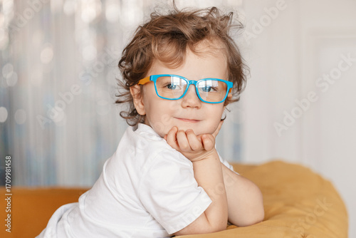 A close-up portrait of a little boy wearing eyeglasses with blue frames, looking cheerful and curious