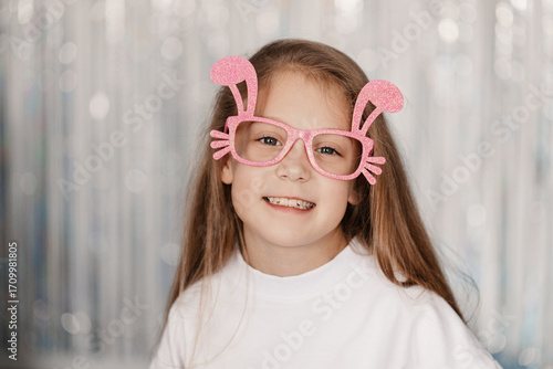 A little girl wearing festive bunny-ear glasses smiles joyfully, surrounded by bright Easter decorations and a cheerful springtime atmosphere