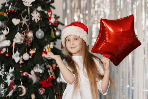 Cheerful 9-year-old girl in Santa Claus hat holding foil balloon shaped like candy cane in front of decorated Christmas tree, festive holiday mood