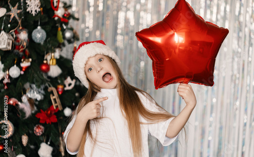 Cheerful 9-year-old girl in Santa Claus hat holding foil balloon shaped like candy cane in front of decorated Christmas tree, festive holiday mood
