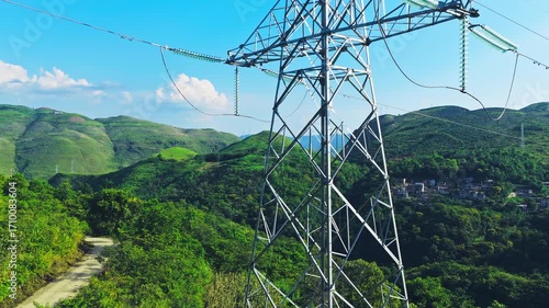 Aerial shot of a high voltage electricity transmission tower and power lines set against a backdrop of lush green rolling hills.