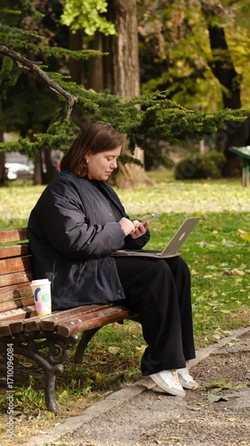 Woman sitting on a park bench with a laptop, using her smartphone and a takeaway coffee beside her. Autumn work and relaxation outdoors. Digital nomad concept. Vertical 4k footage.