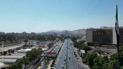 An aerial view captures the new Syrian national flag hoisted on a towering pole in central Damascus, with Mount Qasioun, one of the capital’s most prominent landmarks rising in the background.
