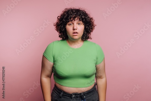 Fotografie Curly-haired individual in green shirt stands against a pink background expressi