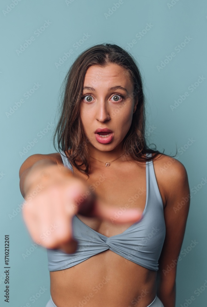 Fototapeta premium Woman looks surprised and points forward against a light blue backdrop indoors during the day