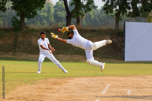 A wicketkeeper in white leaps to catch the ball as a fielder stands ready, on a cricket field