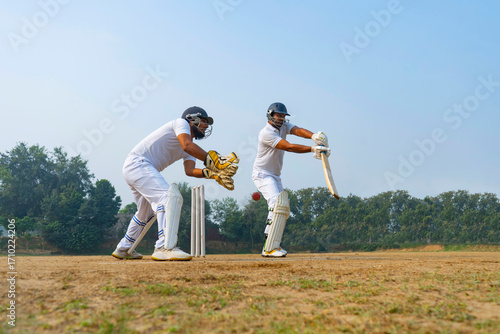 A batsman in white is poised to strike the ball, with a wicketkeeper anticipating the play on a grassy field