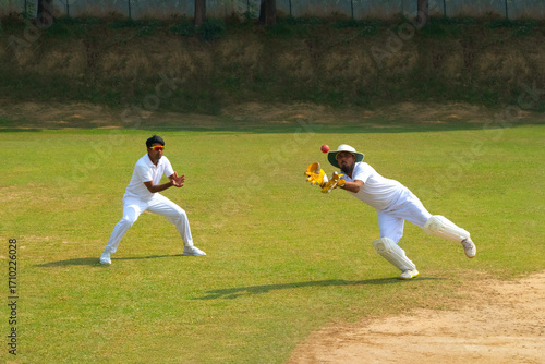 A wicketkeeper catches the ball with a fielder nearby, set on a cricket field under a clear sky, showcasing teamwork