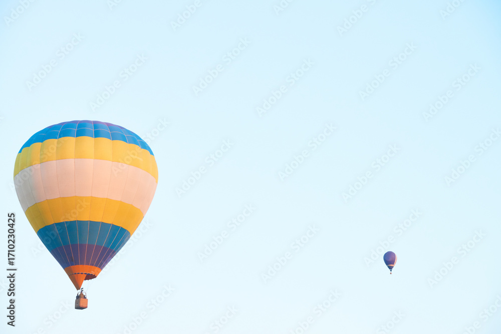 Obraz premium Two hot air balloons floating in clear sky, large striped balloon in foreground carrying people in basket, smaller balloon in distance, empty blue background providing copy space