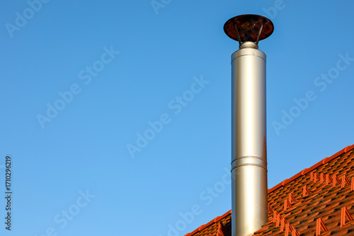  Metal chimney rises from a red tiled roof under a clear blue sky. Sunlight gleams on the polished flue capped with a round cover, symbolizing heating, ventilation, and modern architecture.