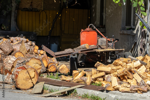 Fotografie A robust log splitter sits amidst large piles of freshly cut and split firewood outside a house, ready for winter heating
