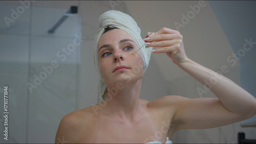 A woman applies serum to her face while looking in a mirror, surrounded by modern bathroom decor and serenity.