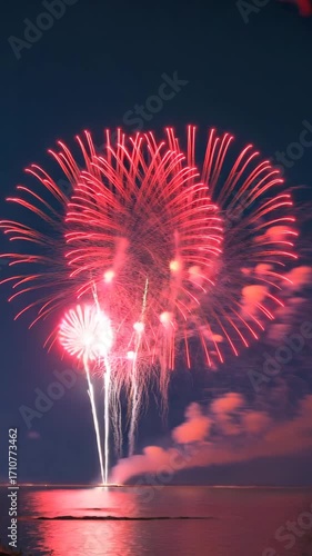 Large red fireworks explode in the night sky with reflection over the sea water.