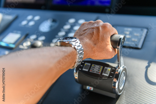 A close-up view of a yacht or ship bridge with a person’s hand operating the throttle controls. The navigation radar screen and electronic control panels are visible in the background