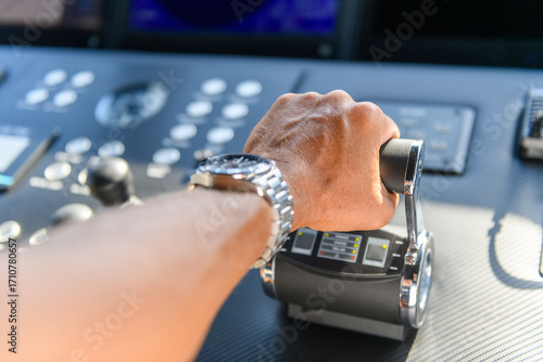 A close-up view of a yacht or ship bridge with a person’s hand operating the throttle controls. The navigation radar screen and electronic control panels are visible in the background
