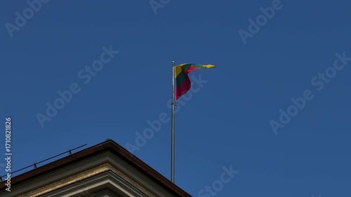 Lithuanian national flag proudly flying against a brilliant deep blue sky, featuring the distinctive yellow, green, and red horizontal stripes on a golden flagpole above a historic building with tradi