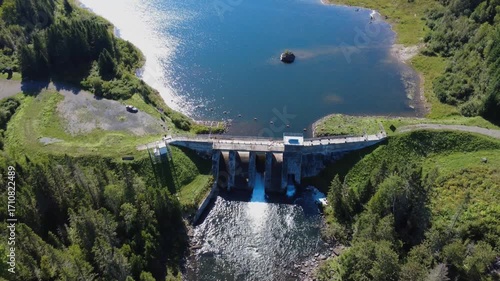 Aerial view of the sparkling Lac Mitis outlet and its old hydroelectric dam where a car is parked in the heart of the mountainous forest. Lac-des-eaux-mortes, Quebec, Canada, 2025.