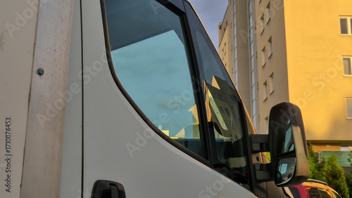 Close-up view of a white van parked near residential buildings, showcasing its side window and mirror. Captures a modern urban environment during the golden hour.