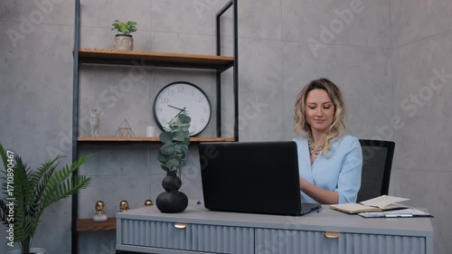 mobile phone, professional attire, corporate lifestyle. In contemporary office, cheerful businesswoman dressed in light blue suit enjoys pleasant phone conversation.