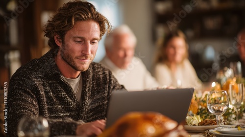 A man using his laptop at the dinner table before the Thanksgiving meal, with family members chatting in the background. Reflects the blend of modern life and tradition.