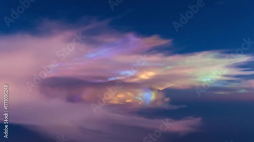 A stunning display of cloud iridescence with shimmering rainbow colors glowing within a wispy pink cloud against a dark blue sky.