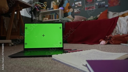 A laptop with a green screen and tracking markers is on a floor, likely a bedroom, with an open notebook nearby. In the background, a bed is visible, covered in a red blanket.