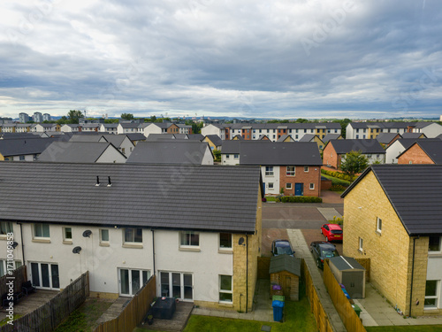 A residential area, featuring a street with houses and trees under a partly cloudy sky, residential street with terraced, semi-detached houses. The image captures an elevated perspective. 