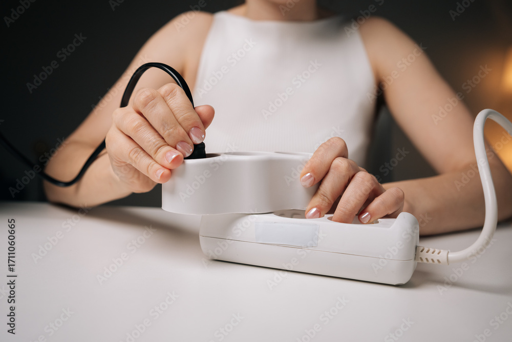 Fototapeta premium Close-up cropped shot of woman plugging black cable into white power strip on white table, with blurred background and soft lighting, creating sense of carefulness and precision.