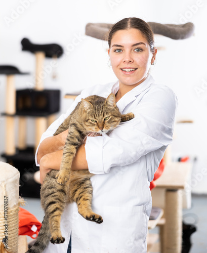 Fotografie Cheerful young female volunteer standing in white coat in animals shelter, holding big gray tabby tomcat found on street