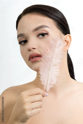 Beautiful young woman with perfect fresh skin and makeup touches her face by feather. Portrait of model with make-up, eyebrows and long eyelashes. Beauty and Spa, skincare and wellness.