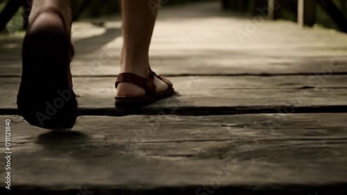 A low-angle shot of a person's feet in sandals walking on a wooden bridge in a forest.