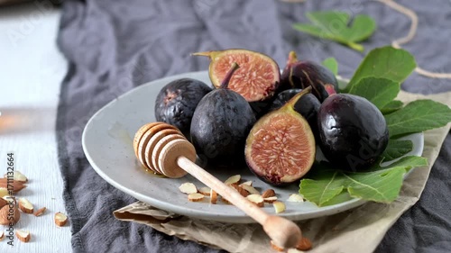 A rustic still life of fresh Black Mission figs, some whole and some cut in half to reveal their vibrant red flesh. The figs are arranged on a gray ceramic plate with a wooden honey dipper and chopped