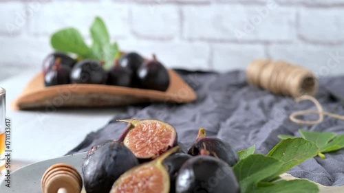A rustic still life of fresh Black Mission figs, some whole and some cut in half to reveal their vibrant red flesh. The figs are arranged on a gray ceramic plate with a wooden honey dipper and chopped