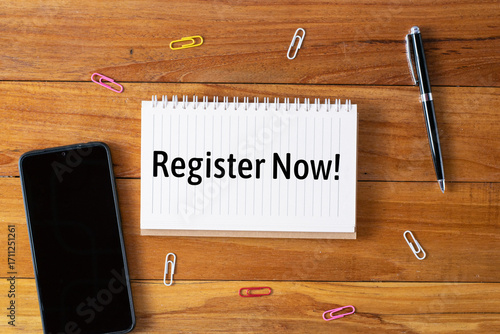 Register now written in a notebook on wooden desk with smartpone, pen and paperclip