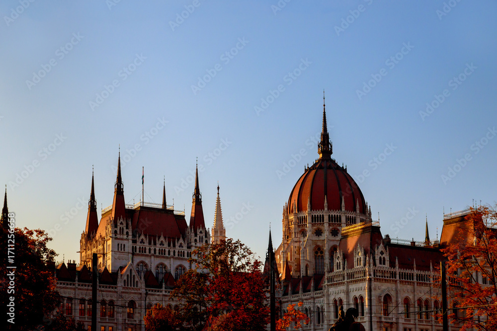 Fototapeta premium Hungarian Parliament Building stands majestically against clear sky during sunset, surrounded by autumn colors.