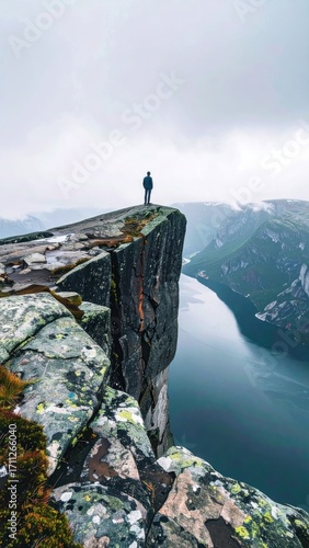 Person standing on a cliff edge with a river flowing between two mountains beneath a cloudy sky on a wet day