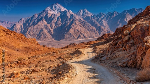 A dusty road leads toward majestic mountains under a clear blue sky, creating a landscape of rugged beauty in the arid region