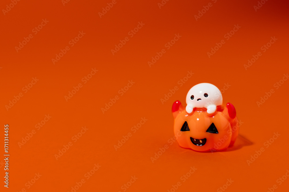 Fototapeta premium playful ghost figurine sitting inside a smiling jack-o’-lantern pumpkin, lit against a bright orange background. The Halloween-themed decoration casts a dramatic shadow