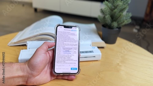 Closeup of hand using smartphone with print preview app beside portable document scanner and book on desk. Great for business, education, technology, and productivity themes.